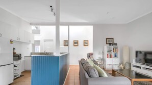 A modern kitchen with white cabinets, a blue island, and a dining area with a grey sofa and a TV. - Cosy Getaway with Fireplace and Spacious Backyard (Broulee)