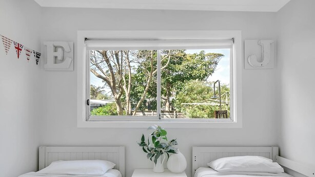 A bedroom with two beds, a window with a view of trees and a bridge, and a small table with a vase of flowers. - Cosy Getaway with Fireplace and Spacious Backyard (Broulee)