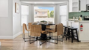 A modern kitchen with a dining area, featuring a round table with chairs, white cabinetry, and a view of a house through the window. - The Palm & Putt: Heated Pool | Golden Tee | Bocce (Phoenix)