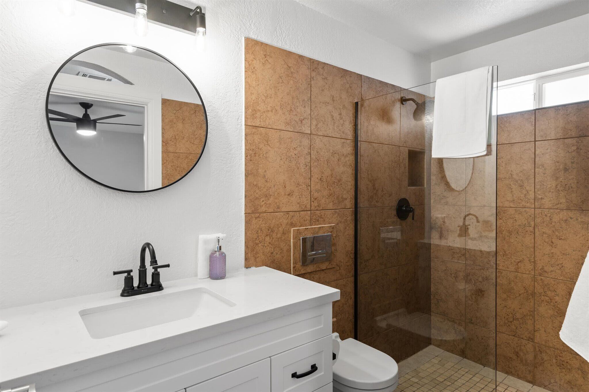 A bathroom with a white vanity, a round mirror, a shower with a glass door, and a white toilet.