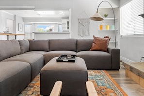 Un salon moderne avec un canapé d’angle gris, un petit pouf, une lampe sur pied et un tapis coloré.