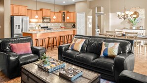 A modern living room with a black leather sofa, a coffee table with decorative items, and a kitchen area with wooden cabinets and stainless steel appliances. - Backyard Oasis | Pool, Theater & Fire Pit (Glendale)