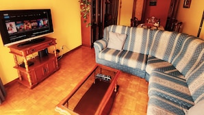 Un salon avec un téléviseur posé sur un meuble en bois, un canapé bleu et blanc à motifs, une table basse en bois et un lustre.