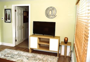 A living room with a television on a wooden stand, a decorative '@' sign on the wall, and a small table with a vase.