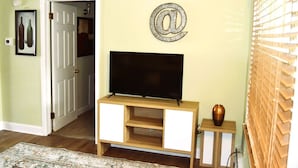 Ein Wohnzimmer mit Fernseher auf einem Holzboden, einem dekorativen '@' Schild an der Wand und einem kleinen Tisch mit einer Vase.