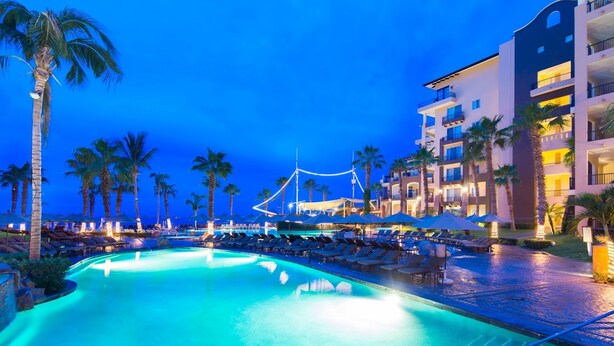 A swimming pool with lounge chairs and umbrellas, surrounded by palm trees and a hotel building.