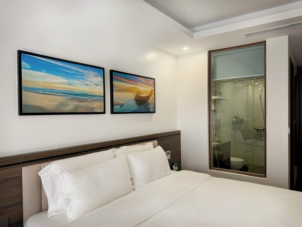 Habitación de hotel con cama, dos cuadros en la pared y un baño visible a través de una puerta de vidrio.
