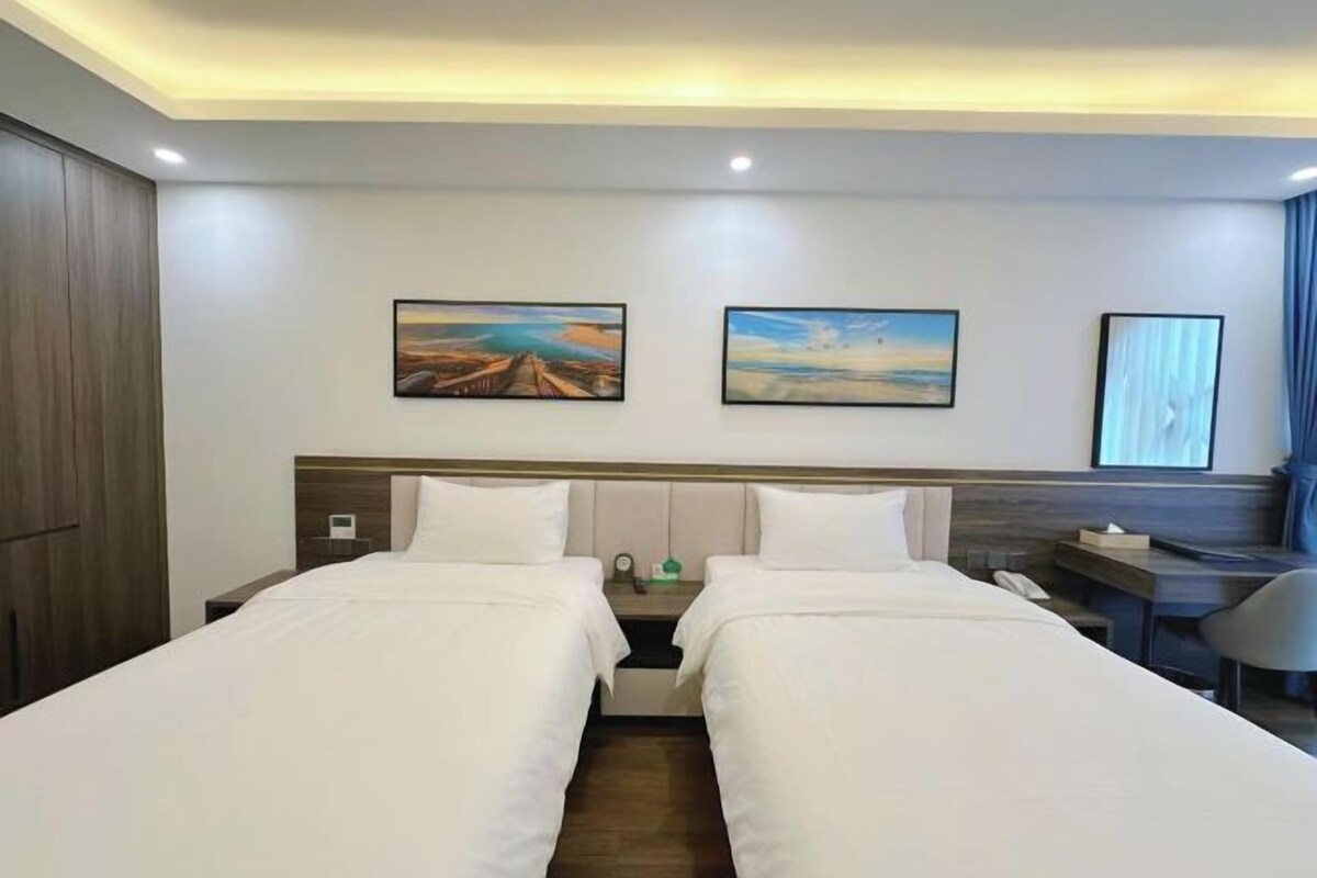 Habitación de hotel con dos camas, un escritorio y una silla. Hay dos cuadros en la pared y una ventana con cortinas.
