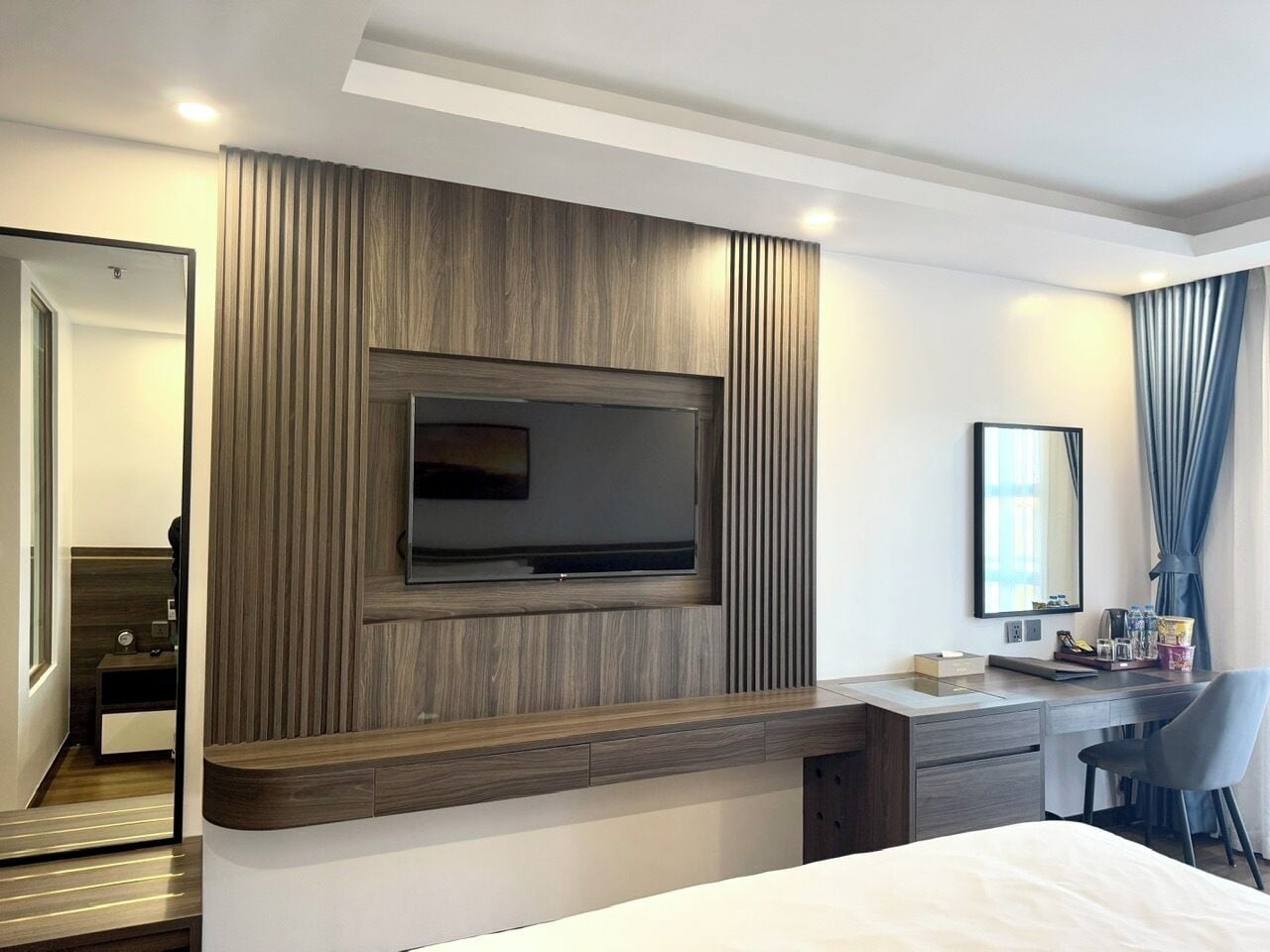 A modern hotel room with a large flat-screen TV mounted on a wooden panel wall, a desk with a chair, a bedside table, and a mirror.