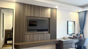 A modern hotel room with a large flat-screen TV mounted on a wooden panel wall, a desk with a chair, a bedside table, and a mirror.