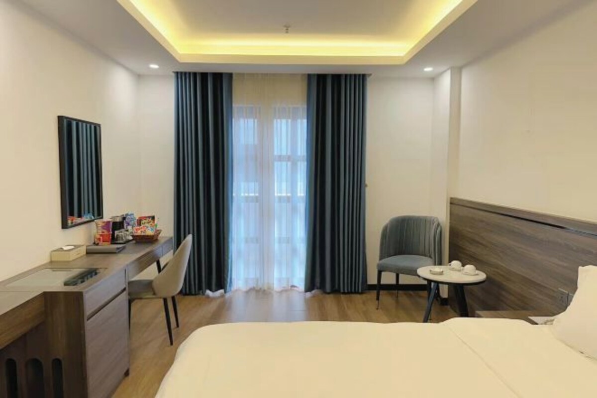 Habitación de hotel con cama, escritorio con silla, una mesita con tazas y ventana con cortinas.
