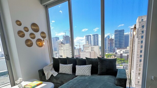 Une chambre moderne avec un grand lit, une petite table blanche et une vue sur les toits de la ville, visible à travers de grandes fenêtres.