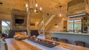 A rustic wooden interior with a stone fireplace, a wooden dining table with bread and decorative items, a kitchen with stainless steel appliances, and a staircase. - Private Indoor Pool + Hot Tub + Sauna + Theater + Game Room | Mins to Gatlinburg (Gatlinburg)