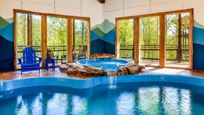 A pool with a waterfall feature, surrounded by wooden deck chairs and tables, with large windows offering views of greenery. - Private Indoor Pool + Hot Tub + Sauna + Theater + Game Room | Mins to Gatlinburg (Gatlinburg)