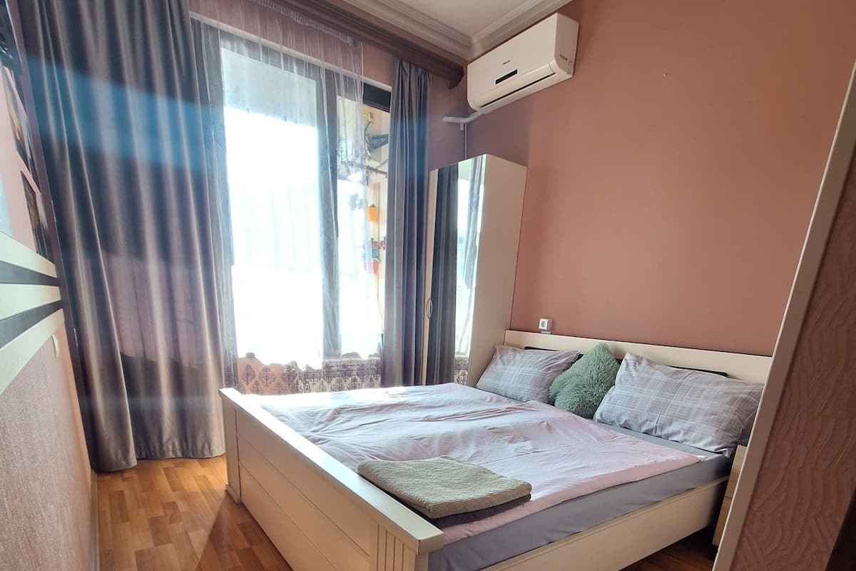A bedroom with a bed, wooden flooring, an air conditioning unit, and a window with curtains.