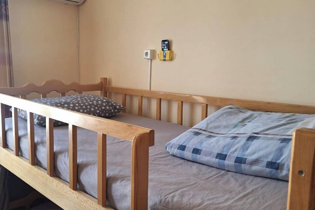 A small, single-bed room with a wooden frame, a grey mattress, and a checkered pillow. There is a wall-mounted air conditioning unit and a power outlet with a phone and a small box.