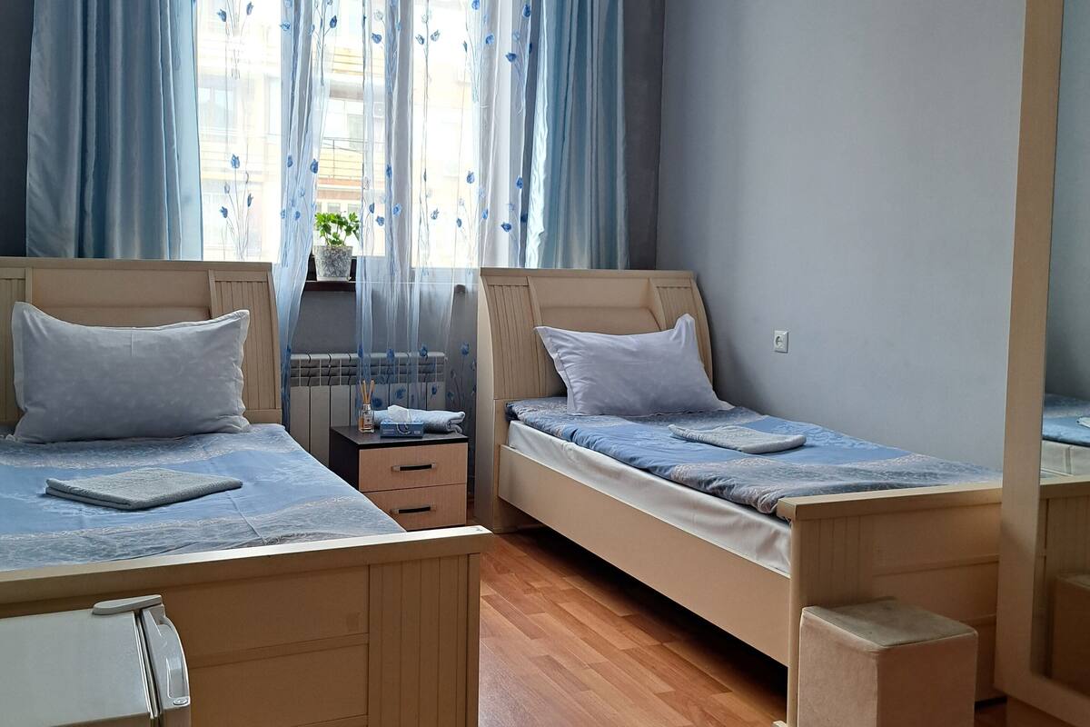 A bedroom with two single beds, a wooden floor, a window with curtains, and a hanging light fixture.