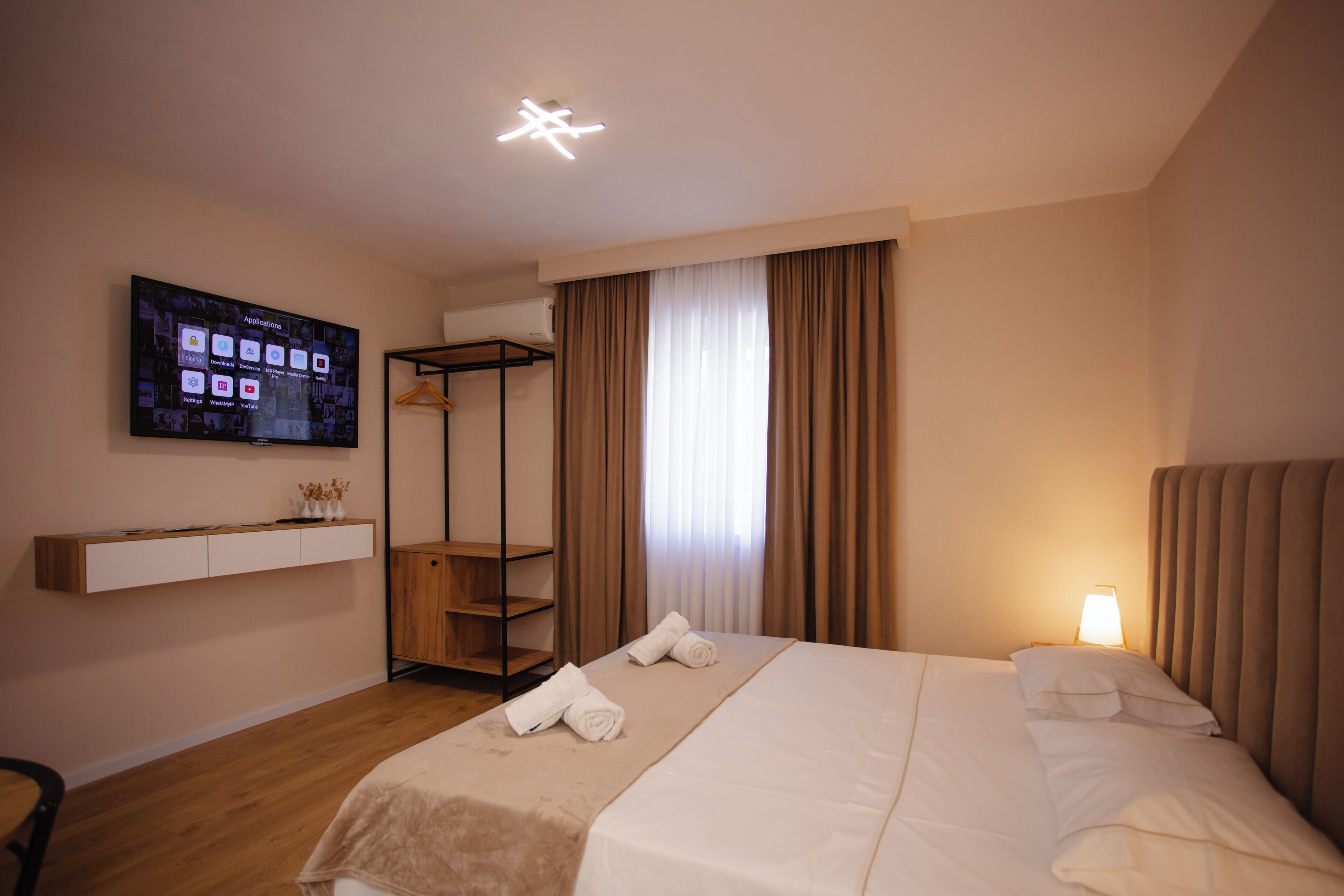 A hotel room with a bed, a TV mounted on the wall, a shelf with decorative items, and a window with curtains.