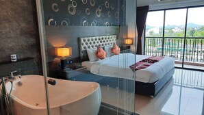 A modern hotel room with a large bathtub, a neatly made bed with two pillows, a bedside table with a lamp, and a view of the city through large windows. - Constancy Jomtien Pattaya Hotel (Sattahip)