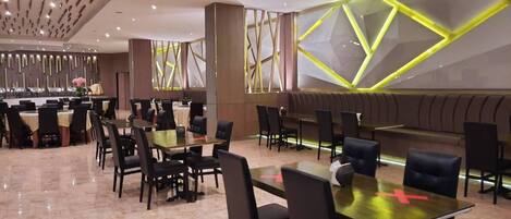 A modern restaurant with multiple dining tables and chairs, a bar area, and a decorative wall with geometric patterns.