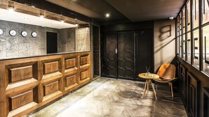 A modern interior with a wooden reception desk, a black door, and a seating area with a round table and chair. - Bellino Hotel (Osan)