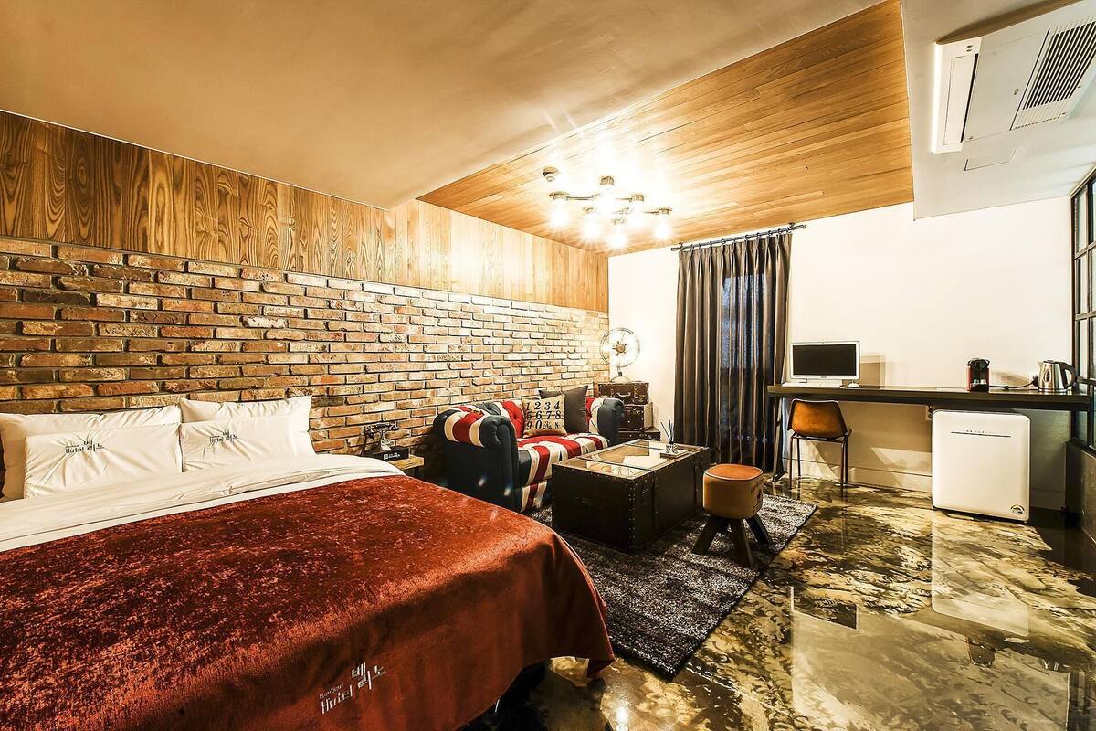 A hotel room with a brick-patterned headboard, a bed with white linens, a red upholstered sofa, a small desk with a computer, and a compact kitchenette.