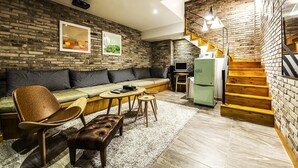 A compact living space with a brick wall, a sofa, a dining area with a table and chairs, a small kitchenette, and a staircase. - Bellino Hotel (Osan)
