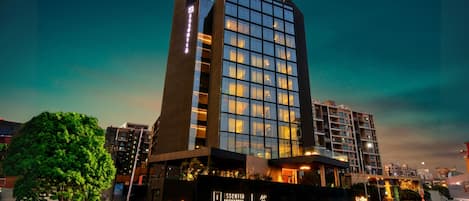 A modern hotel with a prominent glass facade, a well-lit entrance, and a landscaped area with trees.