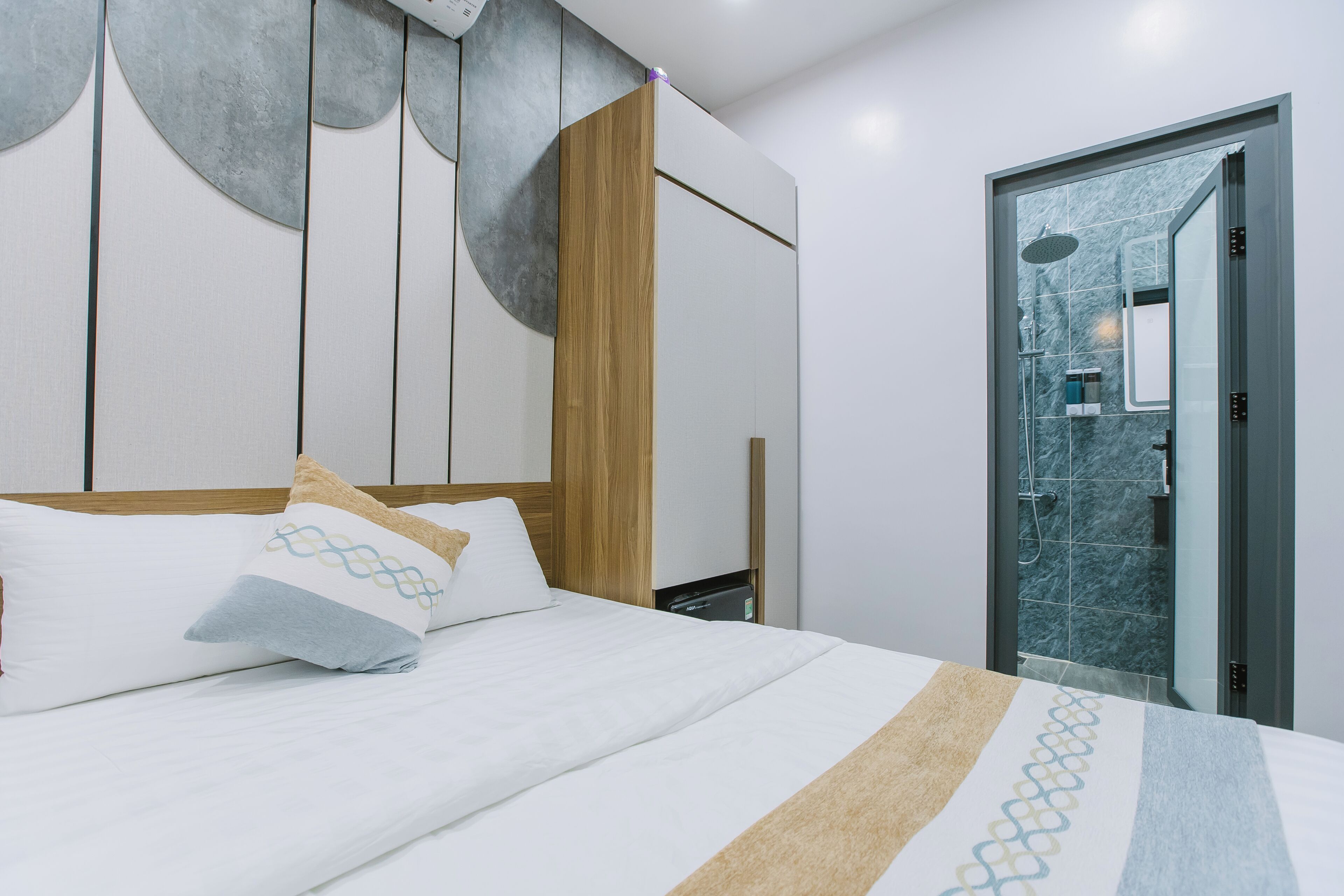Une chambre d’hôtel moderne avec un lit, une tête de lit en bois et une salle de bain visible à travers une porte en verre.