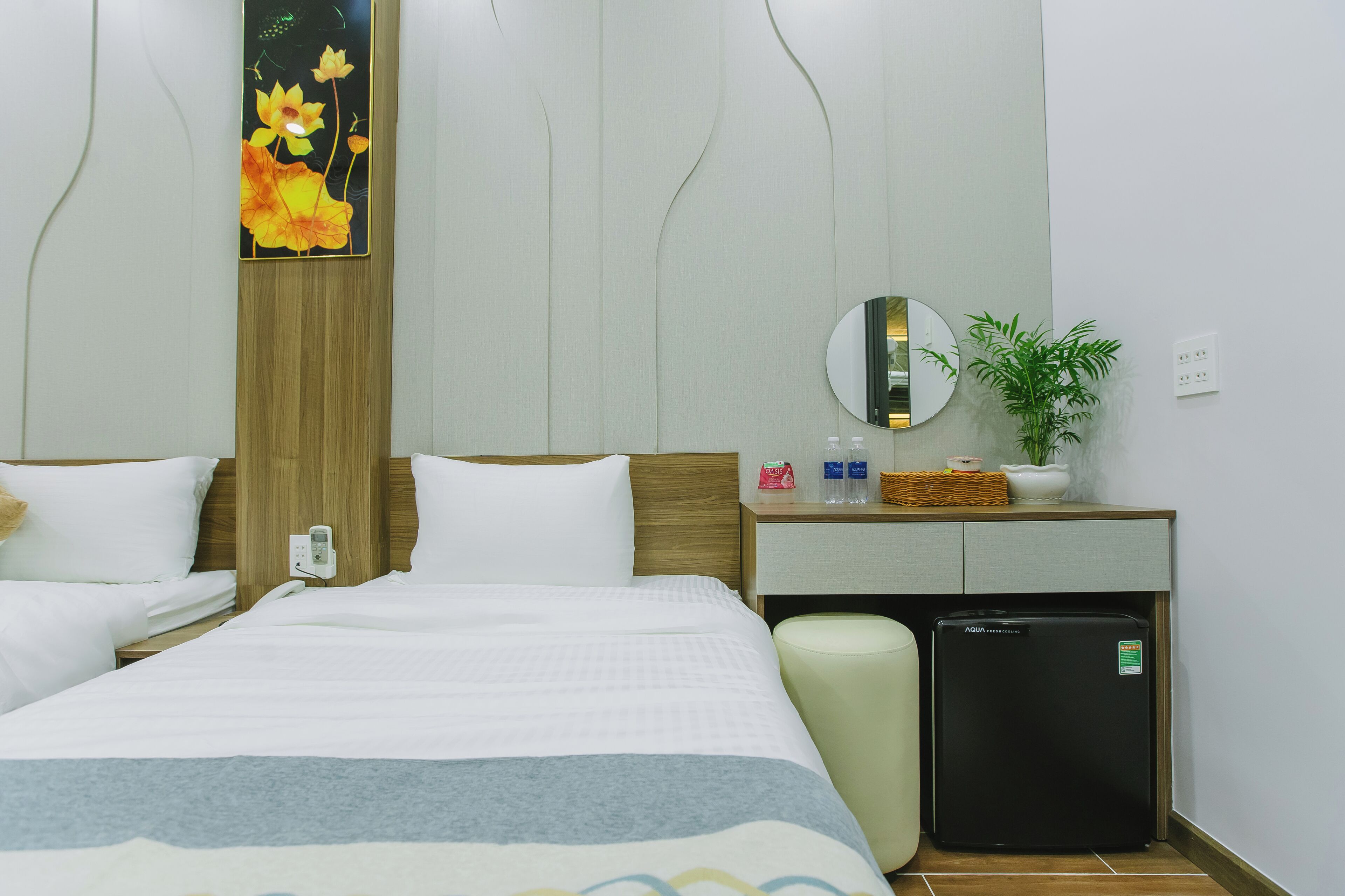 Quarto de hotel com cama, mini-geladeira, mesa de cabeceira com espelho e planta na parede.