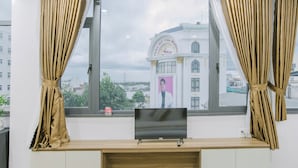 A room with a large window, curtains, a flat-screen TV, and a view of a building with a sign. - Peony Hotel (Can Tho)