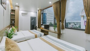 A hotel room with two beds, a desk with a computer, a window with curtains, and a view of a cityscape. - Peony Hotel (Can Tho)