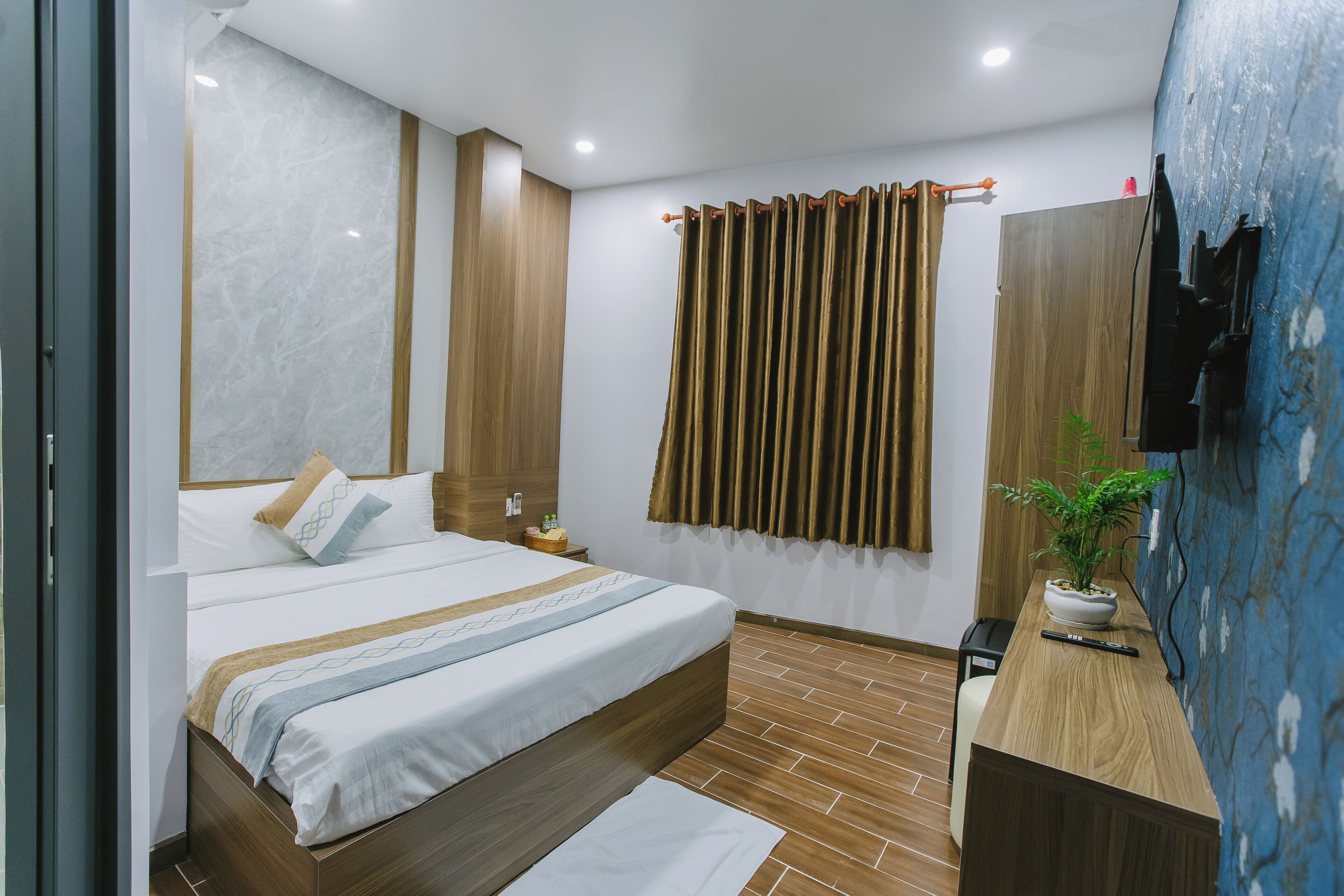 Een moderne slaapkamer met een groot bed, een houten hoofdbord, een klein nachtkastje, een nachtlampje, een potplant en een tv aan de muur.