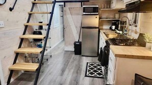 Une cuisine compacte équipée d’un four à micro-ondes, d’un réfrigérateur et d’une cuisinière intégrés. Une échelle en bois et des étagères sont fixées au mur, et un petit tapis est posé sur le sol.
