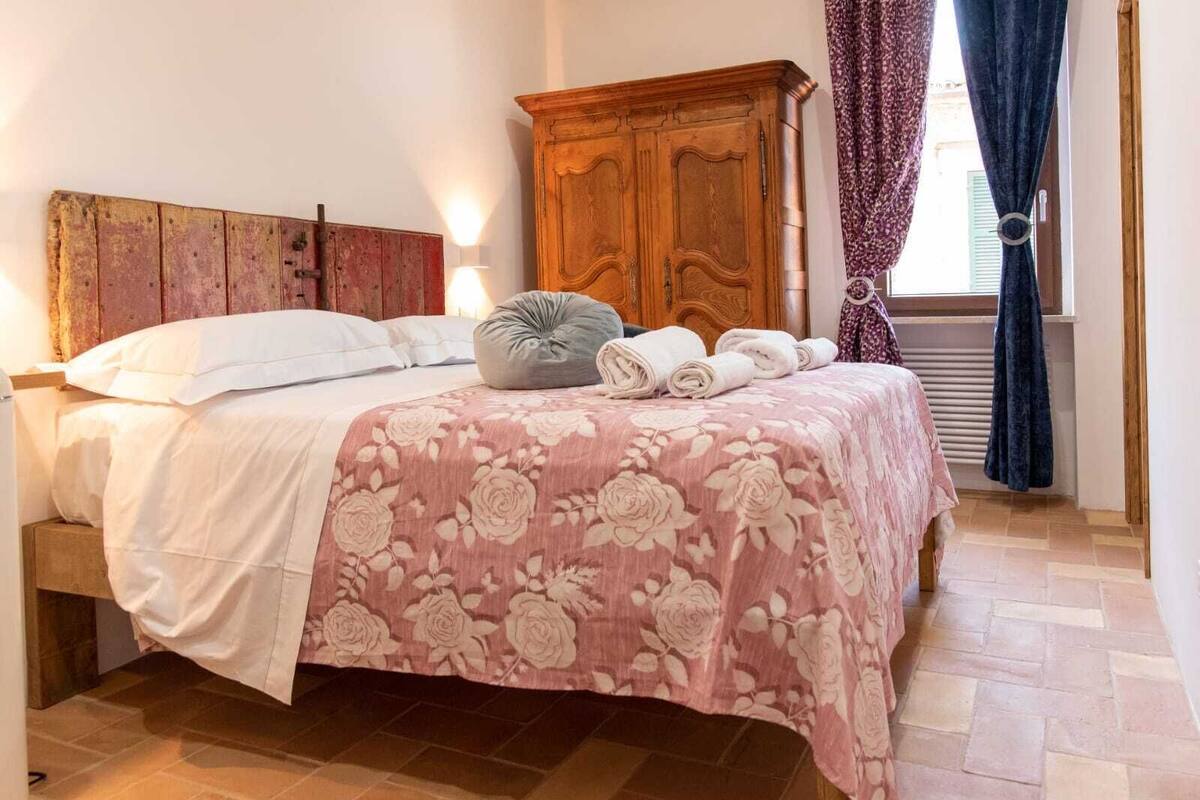 Un dormitorio con cabecera de madera, una cama con colcha estampada y una almohada gris, además de toallas enrolladas sobre la cama.