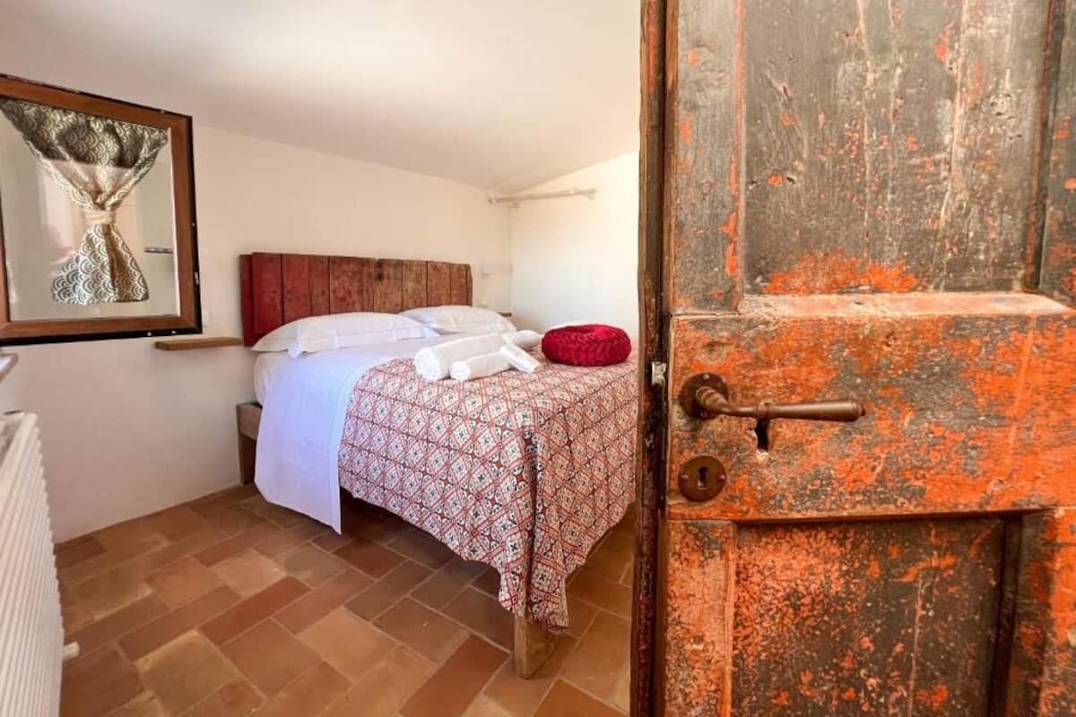 Un dormitorio con una cama, un almohadón rojo, un cabecero de madera y una puerta rústica.