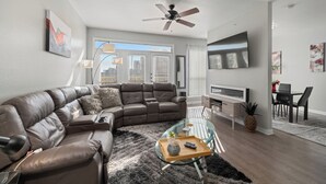 A modern living room with a sectional sofa, a glass coffee table, a ceiling fan, a TV, and a dining area. - Spacious Dallas Apartment with City Views Near Galleria Mall (Dallas)