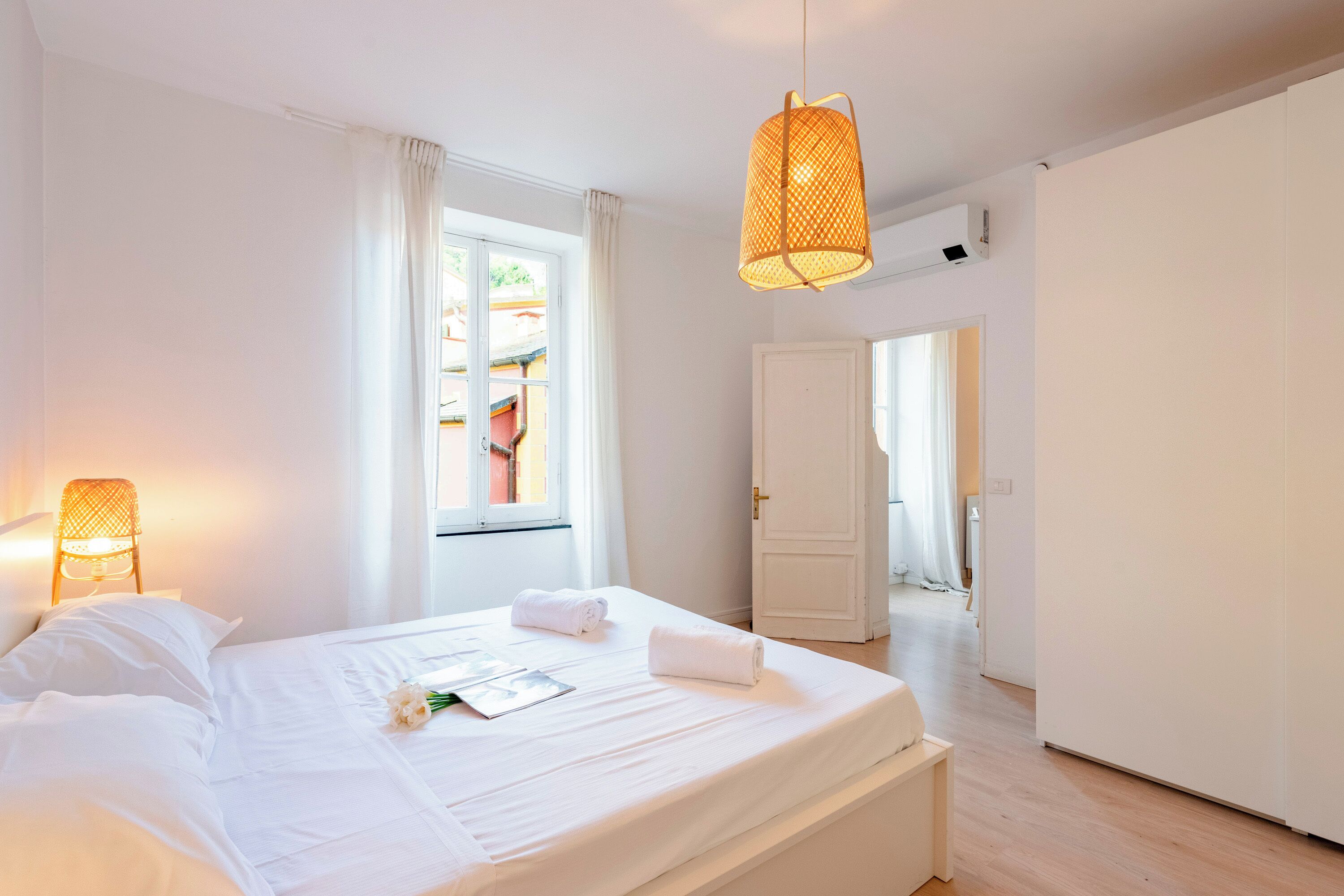 A hotel room with a large bed, a window with curtains, a hanging light fixture, and a door leading to another room.