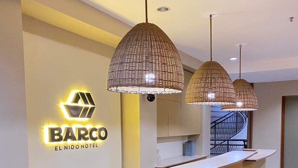 A hotel reception area with a modern design, featuring a reception desk, two hanging pendant lights, and a staircase in the background.
