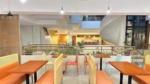 A modern restaurant with wooden tables and orange cushioned benches, a kitchen area in the background, and a staircase leading to an upper level.