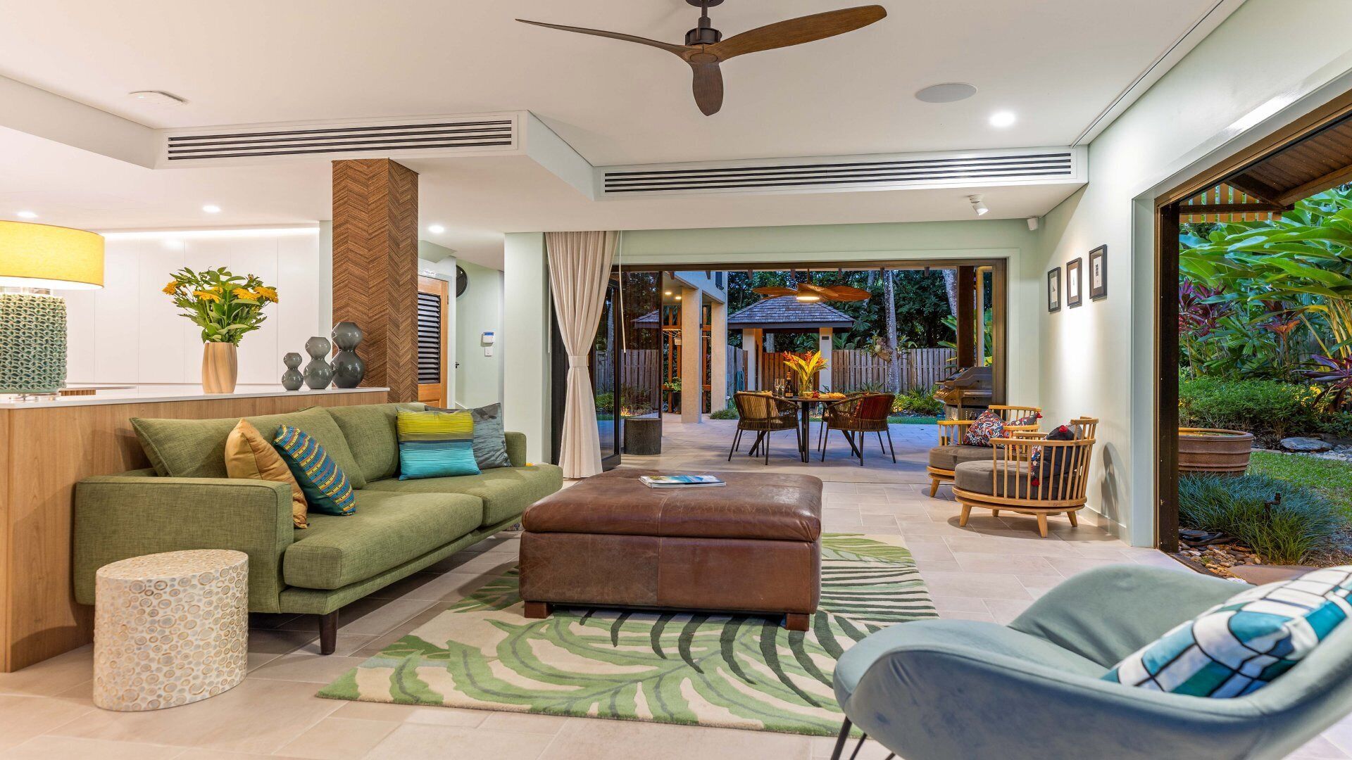 A modern living room with a sofa, ottoman, and chairs, a dining area with a table and chairs, and a view of the backyard with plants and a barbecue.