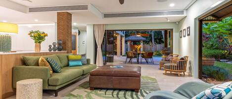 A modern living room with a sofa, ottoman, and chairs, a dining area with a table and chairs, and a view of the backyard with plants and a barbecue.