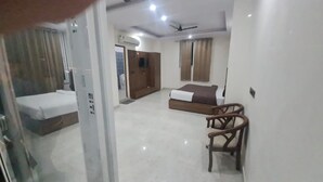 A hotel room with a bed, a chair, a ceiling fan, and a window with curtains. - JSR Hotels (Narendranagar)
