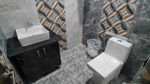 A bathroom with a white sink, a toilet, and a shower area. The walls are tiled with a geometric pattern. - JSR Hotels (Narendranagar)