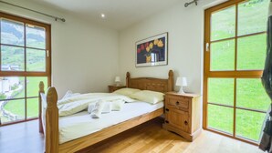 A bedroom with a wooden bed, two bedside tables with lamps, a painting on the wall, and large windows with a view of greenery. - Modern Mountain Apart 5 - on skiing slope (Rauris)