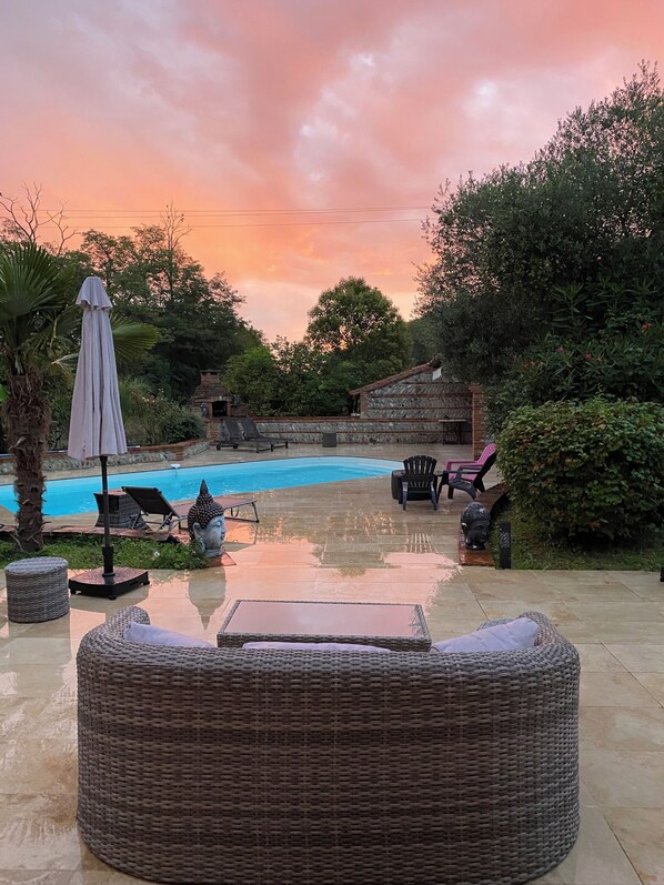Relax by the serene pool surrounded by lush greenery and a stone wall, with comfortable lounge chairs and a wicker sofa.