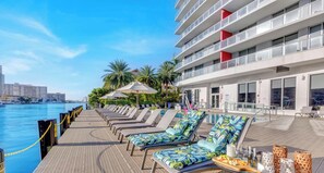 A poolside area with lounge chairs, umbrellas, and a view of the water and buildings.