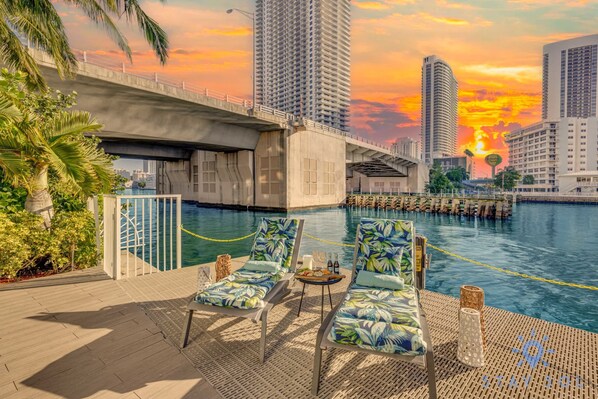 A rooftop pool area with lounge chairs, a table with drinks, and a view of the sunset over a cityscape with tall buildings.