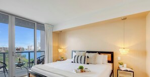 A hotel room with a large bed, two bedside lamps, a nightstand, a small table with a plant, and a view of the city through large windows. - Spectacular Resort, Prime Location, Balcony, Gym (Hallandale Beach)