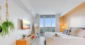 A modern hotel room with a bed, a desk, a chair, a plant, and a large window with a city view. - Spectacular Resort, Prime Location, Balcony, Gym (Hallandale Beach)